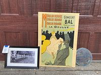 Photo showing a framed black and white photo of a bench, vintage reproduction of Toulouse-Lautrec's Moulin Rouge poster mounted on wood slab, and a round stained glass panel on the ground.