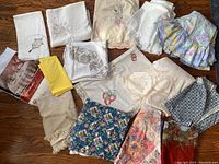 Overview of all vintage tablecloths laid out on a wooden floor, showing variety in patterns, fabrics and colors including embroidery, lace, and floral prints.