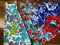 Four vintage Hawaiian clothing items laid out: floral textured white dress, blue hibiscus print dress, blue leaf print dress, and red shirt with printed design and fringe.