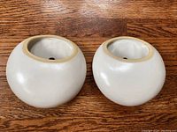 Two white pottery wall pockets shown side by side on a wooden surface, rounded shape with beige rim and visible hole on the back side of each.