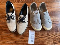 Two pairs of women's shoes on wooden floor. Left pair is Tory Burch black and white oxford-style shoes with black laces. Right pair is Keen light gray slip-on shoes with cross elastic straps and white soles.