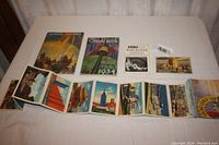 Overview of all ephemera laid out on a cloth surface, showing booklets, postcards, and folder with detailed color images.