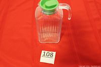 Front view of the clear glass pitcher with olive green plastic lid on a red cloth background. The pitcher is upright, showing its shape and size in relation to the lid.