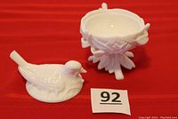 Photo showing the milk glass candy/nut dish with separate lid shaped like a bird sitting on a nest, placed next to the base on a red cloth.