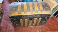 Full frontal of the antique humpback truck trunk on floor showing wood and embossed metal bands on curved lid and front panel.