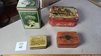 Photo showing all 4 antique metal tins in the lot arranged on a table surface.