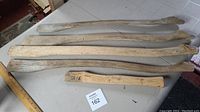 Seven wooden axe handles arranged on table showing variety of shapes, lengths and natural wood colors with signs of age.