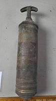 Full view of cylindrical brass fire extinguisher with aged patina and T-shaped handle