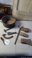 Overview of the entire lot showing a cast iron pot, various axe heads, tongs, and hand tools on a table.