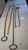 Four older metal fireplace tools on table alongside wooden ruler for scale, including two pokers with loop handles, shovel, and angled tool.