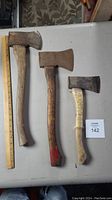 Three used older hatchets of varying size laid on a grey table with a ruler for scale.