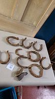 Photo showing nine heavily rusted old horseshoes placed on a white table surface