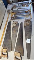 Photo showing 14 vintage hand saws laid out on a table with various blade lengths and shapes. Wooden handles show signs of wear and patina.