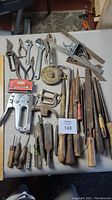 Photo of assorted pliers, wrenches, tin snips, files, a staple gun, wire brush, and putty knife arranged on a white surface.