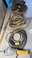 Photo showing the three items in the lot: two coiled extension cords in black and beige colors, and a yellow trouble light with metal cage on a light-colored table.