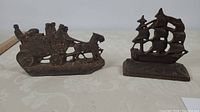 Two cast iron bookends placed side by side on a white surface; one shaped like a stagecoach and horse, the other shaped like a clipper ship with sails.