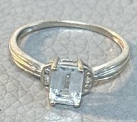 Front close-up of the 10K white gold ring showing the emerald cut clear gemstone and milgrain detailing on band shoulders.