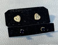 Two pairs of stud earrings displayed on black velvet holders; one pair is heart-shaped gold with floral engraving, the other pair has blue stones in gold prongs.