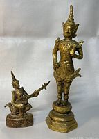 Two Thai brass figurines side by side showing size and detailing; a standing figure in traditional attire and a seated musician playing an instrument.