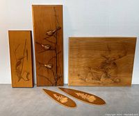 Set of five wooden wall art plaques arranged side by side on a gray surface, showing detailed wildlife carvings including birds and a stag.