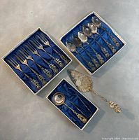 Photo of 7 teaspoons and 5 forks placed in lined boxes with blue cushion, plus a large ornate serving pie/cake server outside the boxes