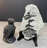 Hand carved dark gray soapstone bird sculpture next to etched soapstone plaque with tree design, both visible in full.