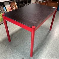 Red and black lacquered dining table with cylindrical legs and rectangular top, photographed assembled on a tile floor.