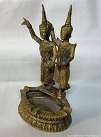 Side angled view of the brass ashtray with two Thai style figurines showing detailed clothing and headpieces, with minor wear visible.