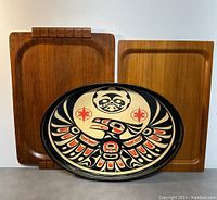 Three trays displayed upright: an oval lithograph print tray with Haida Native Indian art in front, with two wooden trays behind it, one bentwood and one rectangular IKEA tray.