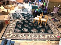 Wide view of the largest and two smaller floral patterned green area rugs with cream borders and fringes on display on basement floor surrounded by household items