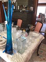 Wide view of dining table covered with clear and colored glass vases, bowls, and plates with tall blue art glass vase dominant in image.