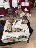 Photo showing three Kachina dolls posed on a woven textile mat with a book titled 'Hopi Kachinas' and a rectangular plaque nearby.