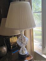 Full view of one crystal table lamp with beige pleated shade near window, showing height and shape.