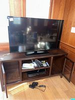 Samsung 46-inch TV model UN46EH5000F on wooden TV stand with glass cabinets, remote controls and other accessories on shelf.