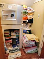 Wide angled photo showing stacked assorted folded towels on shelves and floor with household safety items on floor in front of a small built-in safe and shelving unit.