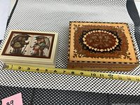 Two music boxes side by side on a patterned surface, showing their relative size with a measuring tape indicating length approx 7-14 inches