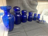Full view of the cobalt blue glass jars, vase, and salt and pepper shaker basket organized on a textured placemat, showing the color and relative sizes of each piece.