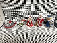 Five Christmas-themed porcelain figurines from left to right: white rocking horse, boy caroler on sled, Mrs. Claus, Santa Claus, and a figure in blue winter clothes holding candle holder.