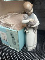 Figurine of a young girl holding a lamb in front of its original blue box with tissue paper inside.