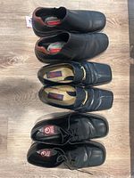 Three pairs of men's black leather dress shoes size 13 arranged on wood floor showing the top view with the Cole Haan slip-on pair, Giorgio S. Bruini loafers with gold bit, and Gran Emyco lace-ups.
