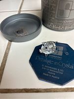 Small crystal pig figurine with one detached ear, blue Swarovski authenticity card underneath, silver metal case lid and base partially visible.