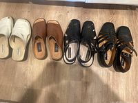 Four pairs of men's leather sandals in size 13 lined up side by side on a wood floor: white slides, brown slip-ons, black slingbacks, black gladiator sandals.