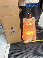 Angel tree topper standing next to its original Avon Gift Collection box, lit up on a black and white surface, shows height with a measuring tape.