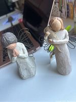 Two WillowTree angel figurines with wire wings, one with folded hands, one holding a bird, posed on a surface near a TV.