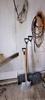 Three shovels of different sizes with black D-shaped handles, placed upright against a wall near the floor with a shovel blade rest.