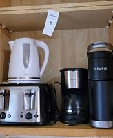 Photo showing Black and Decker two-slot toaster, black Keurig coffee maker, white Sunbeam electric kettle, and Betty Crocker coffee pot on a wooden shelf.