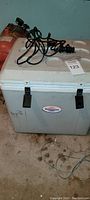 White and gray large electric cooler with coiled power cords on top, placed on a concrete floor with a fire extinguisher visible behind.