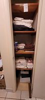 Photo showing the linen closet with folded white and dark gray sheets on the top shelf, assorted towels and lightbulbs on the middle shelves, and storage boxes on the bottom shelf.