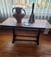 Occasional table with ceramic vase and metal bell on top, placed near window blinds on wooden floor