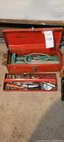 Open red metal toolbox showing assortment of hand tools including pipe wrench, screwdrivers, pliers, and hand saw.